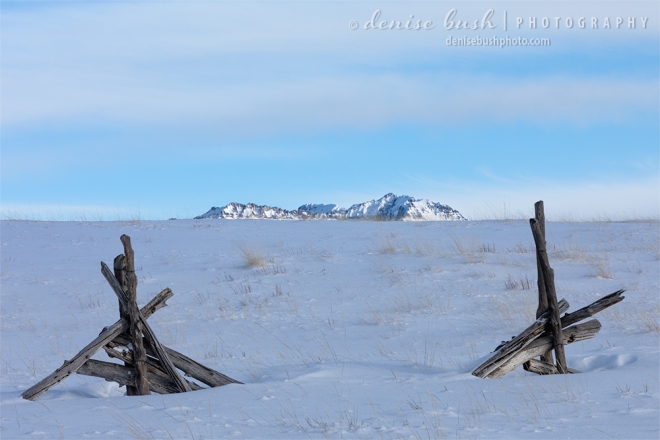 An old broken fence, buried under the snow acts as a frame for the mountain peaks beyond.